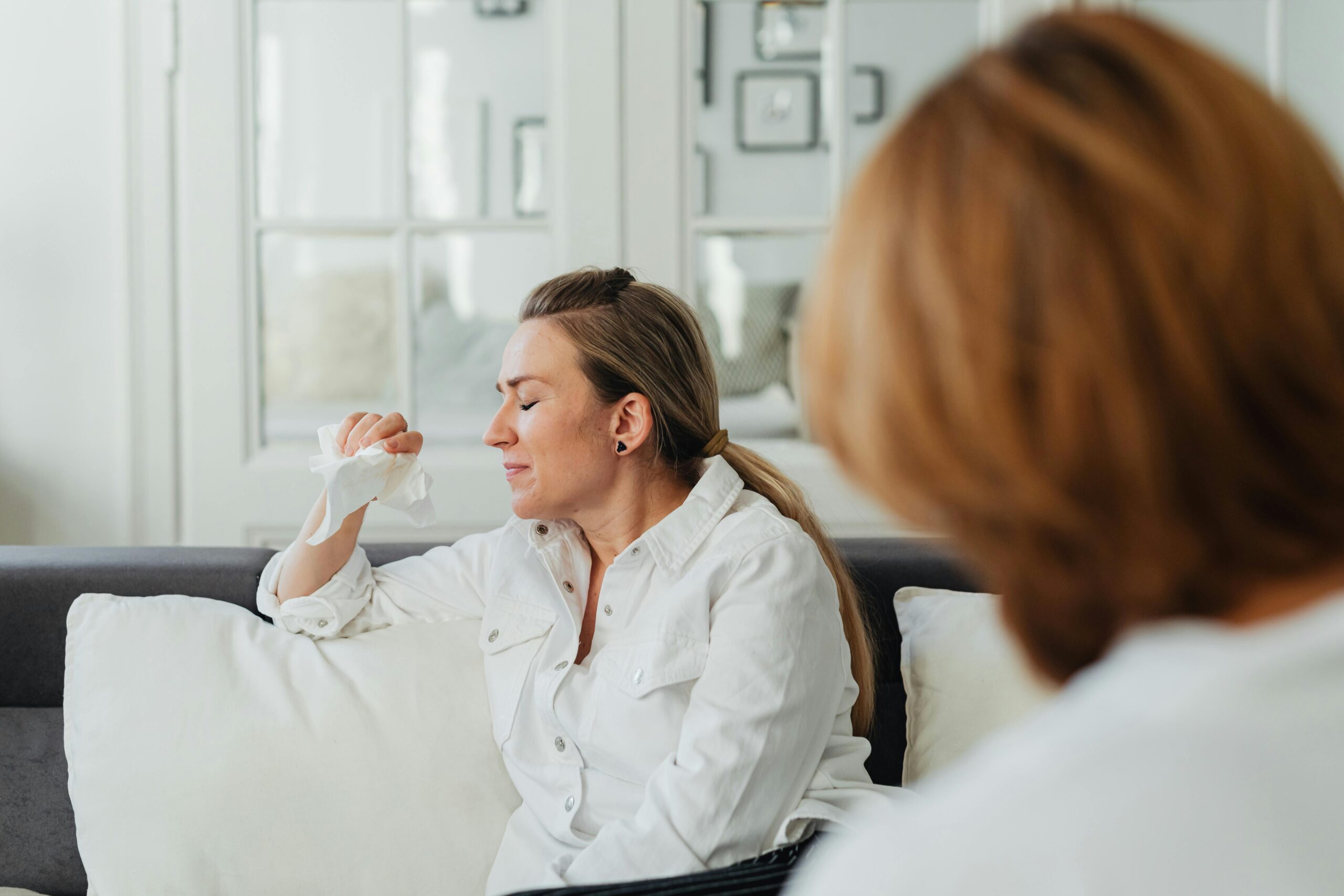 A woman expresses strong emotions on a sofa, holding a tissue indoors.