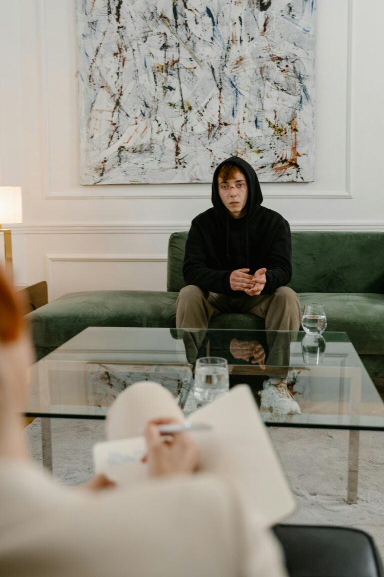 A man in a black hoodie sits on a couch, attending a therapy session indoors.